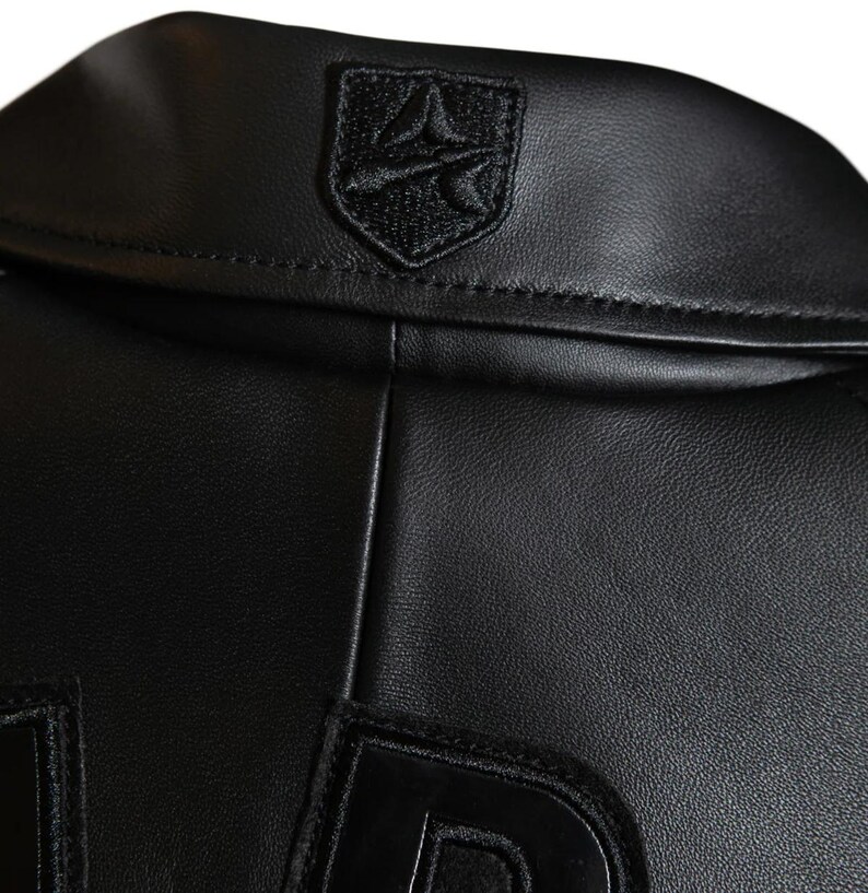 May include: Close-up of a black leather jacket collar with a small, embroidered emblem. The jacket has a classic design, with a smooth, textured surface. The emblem is a shield shape with a stylized design.