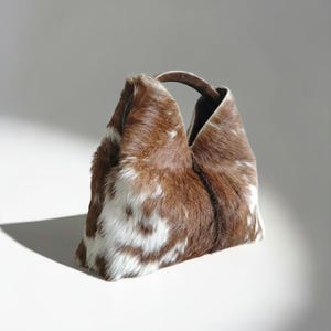 May include: A handbag crafted from brown and white cowhide featuring a textured, fur-like surface. The bag has a structured, triangular silhouette with an integrated top handle and a central seam running vertically down the front panel.