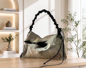 May include: A shoulder bag crafted from textured, multi-tonal gray and black animal hide featuring a distinct, knotted black handle. The bag has a soft, slouchy silhouette with a long, thin black cord accent hanging from the side attachment point.