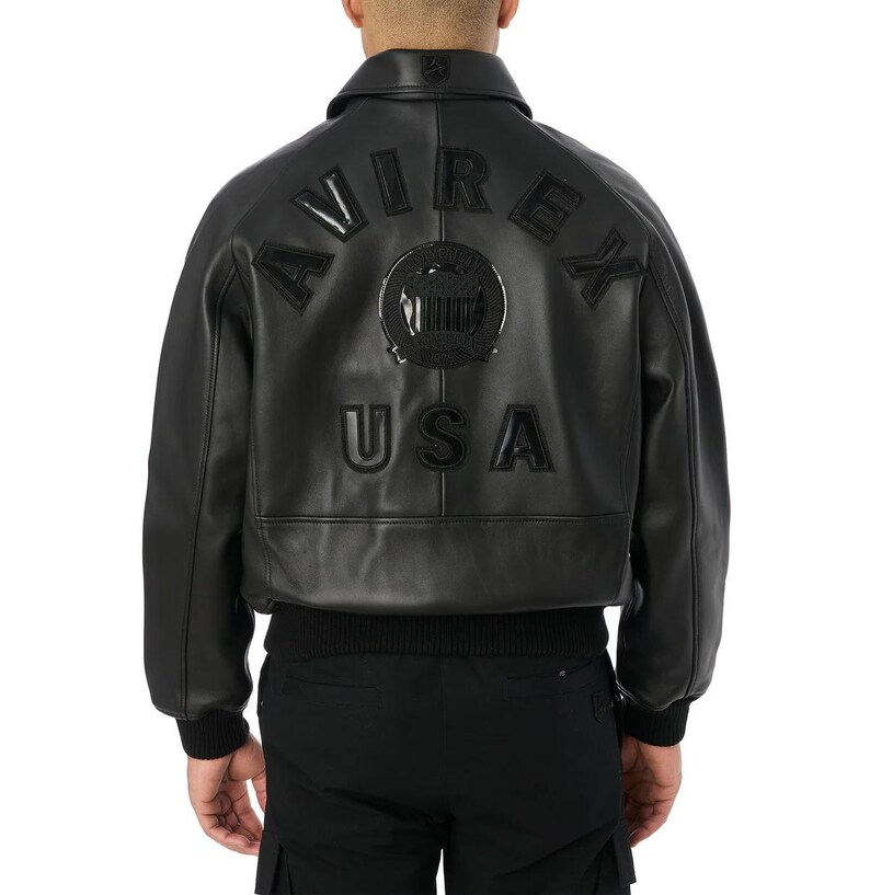 May include: Black leather bomber jacket with the word "AVIREX" and "USA" stitched on the back. The jacket has a ribbed hem and cuffs, a collar, and a zip closure. The jacket is a classic style.