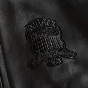 May include: Close-up of a black leather jacket with a textured surface. A black emblem with the text "AVIREX" and "ESTABLISHED NYC 1975" is stitched onto the jacket.