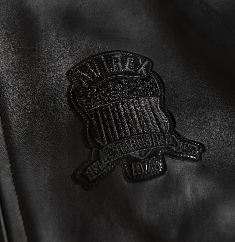 May include: Close-up of a black leather jacket with a textured surface. A black emblem with the text "AVIREX" and "ESTABLISHED NYC 1975" is stitched onto the jacket.