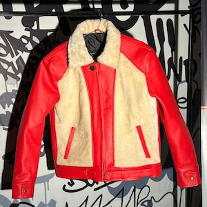 May include: A red and cream-colored jacket with a shearling collar and lining. The jacket features red leather sleeves and panels, a cream-colored shearling interior, and a buckle closure at the waist. The jacket is hanging on a black metal rack.
