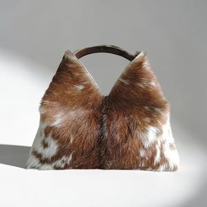 May include: A handbag featuring a brown and white cowhide pattern with a textured fur finish. The bag has a distinct V-shaped top opening and a dark, curved handle integrated into the upper structure, creating a structured silhouette.