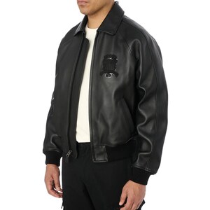 May include: A black leather bomber jacket with a front zipper closure, a collar, and a black emblem on the chest. The jacket has ribbed cuffs and a ribbed hem. The model is wearing a white shirt underneath.