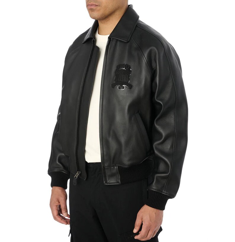 May include: A black leather bomber jacket with a front zipper closure, a collar, and a black emblem on the chest. The jacket has ribbed cuffs and a ribbed hem. The model is wearing a white shirt underneath.