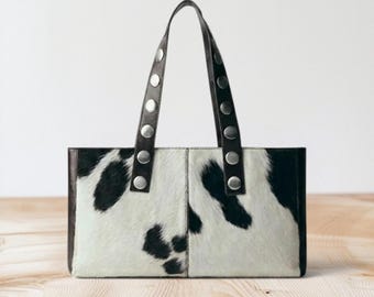 May include: A rectangular handbag featuring a black and white cowhide pattern on the front panels. The bag includes two dark brown leather straps, each adorned with a vertical row of five circular silver-toned metal studs along the length.