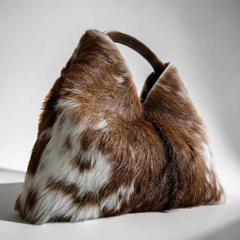 May include: A triangular-shaped handbag crafted from brown and white cowhide with a textured, furry surface. The bag features a single integrated handle at the top, creating a distinct geometric silhouette with a natural, multi-tonal pattern.
