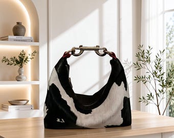 May include: A hobo-style handbag featuring a black and white cow-print pattern with a textured hair-on-hide finish. The bag includes a metallic silver-toned hardware handle attachment with red accents at the connection points and a zipper closure.