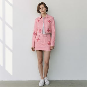 May include: A light pink jacket and matching mini skirt set. The jacket features a collared neckline, long sleeves, and decorative cross-shaped accents. The skirt has a matching cross design. The model is wearing white sneakers.