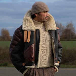 Può includere: Giacca in pelle marrone e nera con colletto e fodera in montone beige. La giacca ha una chiusura a zip e cinturini in pelle marrone. Sono visibili anche un berretto in maglia beige e un maglione a trecce beige.