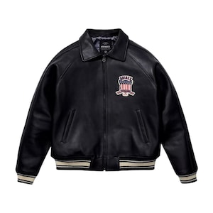 May include: Black leather jacket with a collared neckline and a zip closure. The jacket features a patch with a red, white, and blue design. The cuffs and hem have a striped pattern in white and beige. The brand name "AVIREX" is visible.