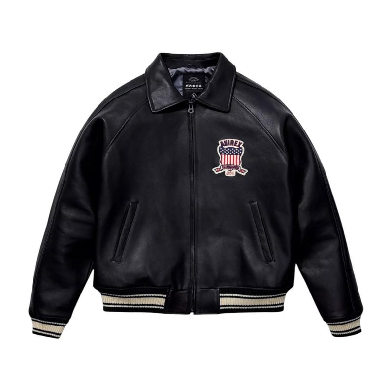 May include: Black leather jacket with a collared neckline and a zip closure. The jacket features a patch with a red, white, and blue design. The cuffs and hem have a striped pattern in white and beige. The brand name "AVIREX" is visible.