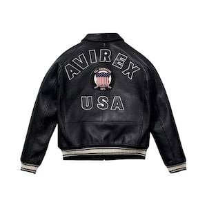 May include: Black leather jacket featuring the text "AVIREX" and "USA" in white lettering. A circular patch with a US flag design is centered between the words. The jacket has a ribbed collar and cuffs with cream and white stripes.