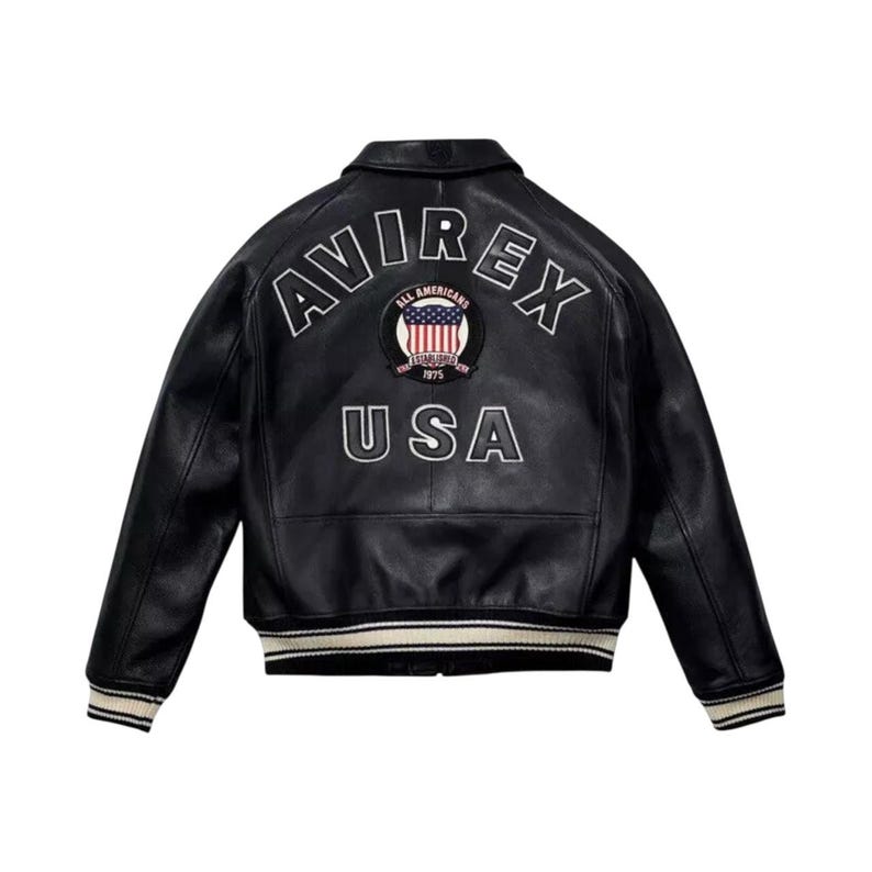 May include: Black leather jacket featuring the text "AVIREX" and "USA" in white lettering. A circular patch with a US flag design is centered between the words. The jacket has a ribbed collar and cuffs with cream and white stripes.