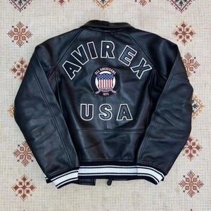 May include: Black leather jacket with white trim on the cuffs and hem. The back displays the word "AVIREX" in white, a circular patch, and the word "USA".