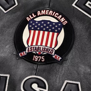 May include: Close-up of a black leather jacket with a circular patch. The patch features the text "ALL AMERICANS" in red and white, surrounding a shield design with the American flag. The text "ESTABLISHED 1975" is below the shield.