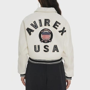 May include: Cream-coloured bomber jacket with black and white striped trim. The back features the word "AVIREX" in large black letters, along with a circular patch with the words "ALL AMERICANS" and "USA".