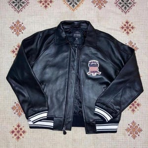 May include: Black leather jacket with a zip fastening, a collar, and a chest patch. The jacket has white stripes on the cuffs and hem. The patch displays "AVIREX" and a design in red, white, and blue colours.