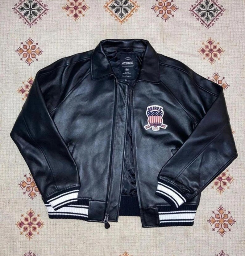 May include: Black leather jacket with a zip fastening, a collar, and a chest patch. The jacket has white stripes on the cuffs and hem. The patch displays "AVIREX" and a design in red, white, and blue colours.