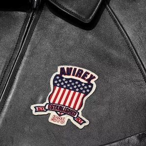 May include: Close-up of a black leather jacket with a colourful patch. The patch features the word "AVIREX" above a shield design with red and white stripes and blue stars. The patch also includes the text "NY ESTABLISHED 1975".