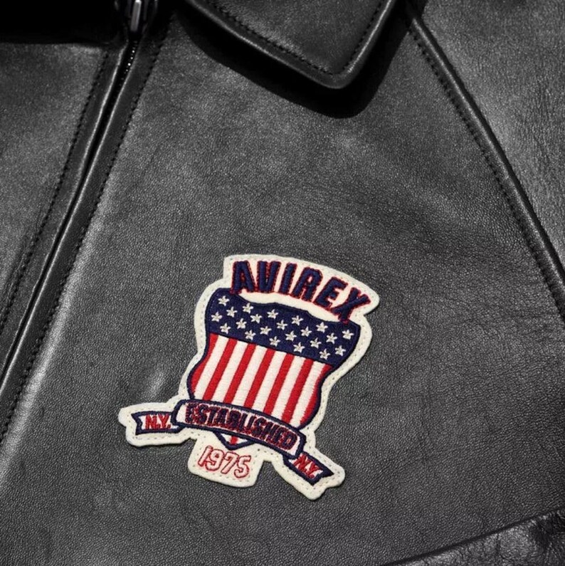 May include: Close-up of a black leather jacket with a colourful patch. The patch features the word "AVIREX" above a shield design with red and white stripes and blue stars. The patch also includes the text "NY ESTABLISHED 1975".