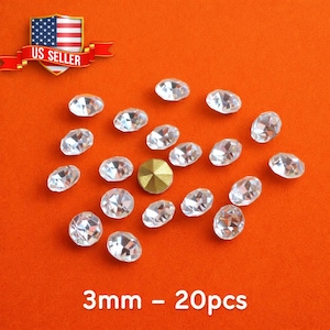May include: Clear, faceted rhinestones and a single gold-colored rhinestone are scattered on an orange surface. The rhinestones are oval and round, with a 3mm size. The text "3mm - 20pcs" is at the bottom.