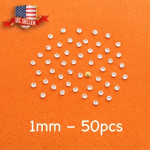 Puede incluir: Pequeños brillantes redondos y transparentes sobre un fondo naranja, con un brillante dorado. El texto "1mm - 50pcs" está en la parte inferior. Una pancarta con la bandera de EE. UU. y "US SELLER" está en la parte superior izquierda. Los brillantes miden 1 mm.