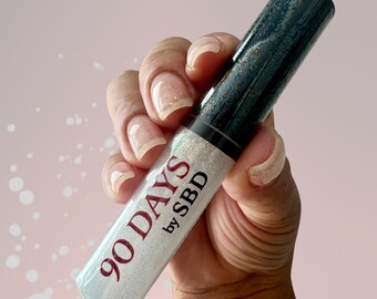 Puede incluir: Un tubo cosmético cilíndrico con una tapa verde oscuro jaspeada y una base transparente rellena de una sustancia brillante que refleja la luz. La etiqueta en la sección transparente presenta el texto 90 DAYS by SBD en color rojo oscuro.