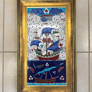 May include: A rectangular wall art piece featuring two ceramic tiles with a hand-painted ship design, framed in a distressed gold-tone border. The artwork displays a large sailing vessel above a smaller boat, rendered in blue, red, and white.