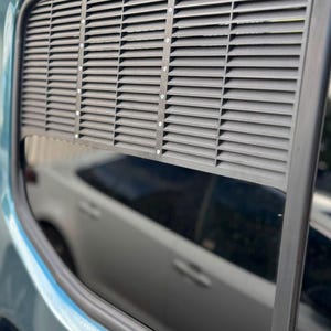 May include: A black louvered vehicle window vent insert installed in the upper portion of a teal car window. The rectangular vent features horizontal slats and a vertical central support bar secured with small silver screws, allowing airflow.