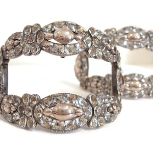 May include: Two antique silver bracelets with clear, sparkling gemstones. Each bracelet features an oval centerpiece with a gold-toned accent, surrounded by smaller, square-cut stones and floral details. The bracelets are displayed on a white background.