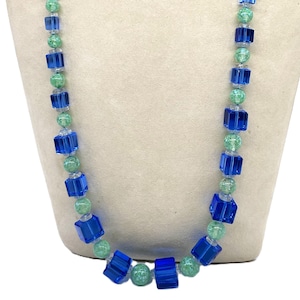 May include: A long necklace made of blue and green glass beads. The beads are square and have a speckled pattern. The necklace is on a white mannequin.