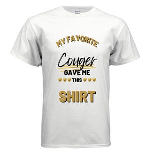 Puede incluir: Camiseta blanca con el texto "MY FAVORITE Couger GAVE ME THIS SHIRT" en letras negras y doradas. La camiseta tiene cuello redondo y mangas cortas.