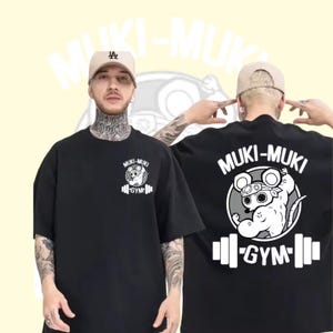 Może przedstawiać: Czarny t-shirt z krótkim rękawem z białym nadrukiem graficznym przedstawiającym umięśnioną mysz w opasce na głowę. Wzór zawiera napis MUKI-MUKI GYM oraz ikonę sztangi. Ten sam motyw w mniejszym rozmiarze znajduje się na lewej stronie klatki piersiowej.