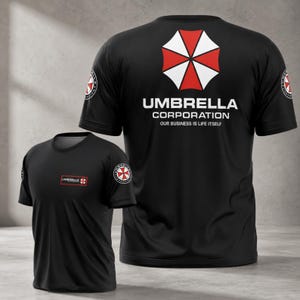 Könnte beinhalten: Schwarze T-Shirts mit dem Umbrella Corporation Logo. Das Logo ist ein rot-weißes Schirmdesign. Der Text "UMBRELLA CORPORATION OUR BUSINESS IS LIFE ITSELF" ist auf der Rückseite aufgedruckt. Ein kleineres Logo befindet sich auf dem linken Ärmel und der Brust.