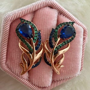 May include: A pair of gold-toned earrings in a feather design, set in a pink velvet jewelry box. Each earring features a large, teardrop-shaped blue gemstone, surrounded by smaller green and blue stones.