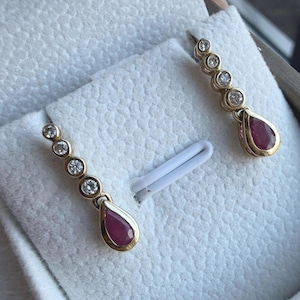 Op de afbeelding: Een paar gouden oorbellen met een druppelvormige robijn en een reeks ronde diamanten. De oorbellen worden gepresenteerd op een wit, getextureerd oppervlak.