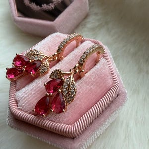 Puede incluir: Un par de pendientes de aro en oro rosa con colgantes en forma de hoja y piedras preciosas rojas en forma de lágrima. Los aros y los detalles en forma de hoja están incrustados con pequeñas piedras transparentes que aportan brillo.