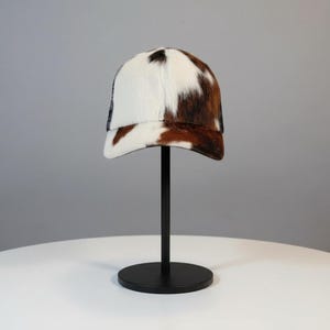 Puede incluir: Gorra de béisbol con estampado de piel de vaca en blanco, marrón y negro. La gorra se exhibe sobre un soporte negro sobre un fondo neutro. La gorra tiene una parte trasera de malla.