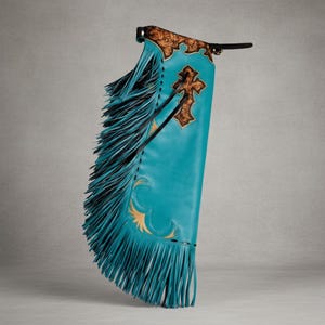 May include: Turquoise leather chaps with brown and gold decorative accents. The chaps feature a cross design and long fringe along the sides and bottom. The top portion has a decorative brown leather design.