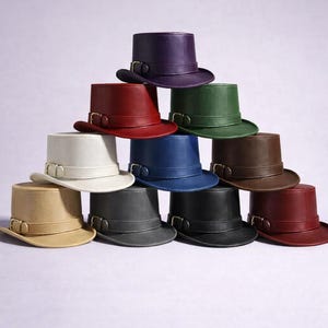 Könnte beinhalten: Eine Sammlung von zehn Leder-Zylindern in verschiedenen Farben, darunter Lila, Rot, Grün, Weiß, Blau, Braun, Beige, Grau, Schwarz und Burgunderrot. Jeder Hut hat ein Band mit einer Schnalle.