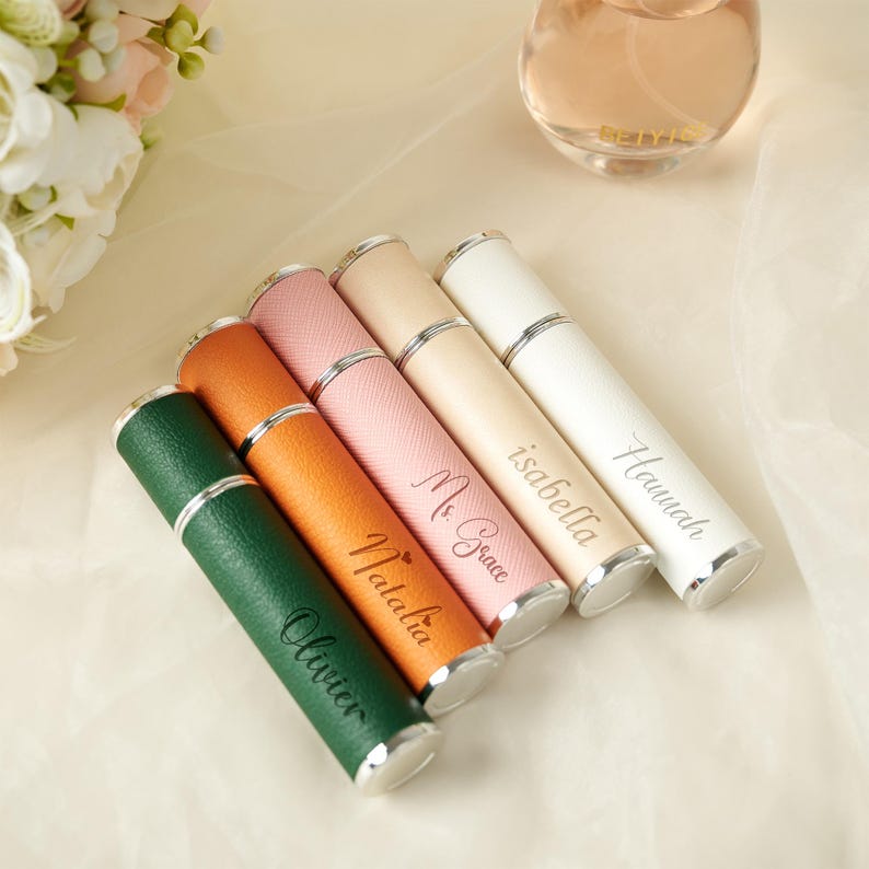 May include: A collection of personalized perfume atomizers in various colors, including green, orange, pink, beige, and white. Each atomizer has a silver cap and is engraved with a name. A perfume bottle is visible in the background.