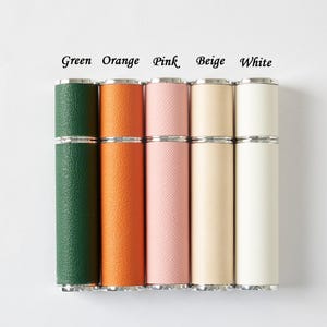 May include: A collection of five cylindrical perfume atomizers in various colors: green, orange, pink, beige, and white. Each atomizer has a silver-colored metal band at the top and bottom. The text "Green Orange Pink Beige White" is above the atomizers.