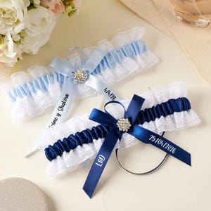 Può includere: Due giarrettiere da sposa con volant in pizzo bianco e nastri di raso in azzurro e blu navy. Ogni giarrettiera presenta una decorazione centrale in strass e nastri di raso stampati con Jaime y Shakira, 4/14/26, L&G e 25/04/2026.