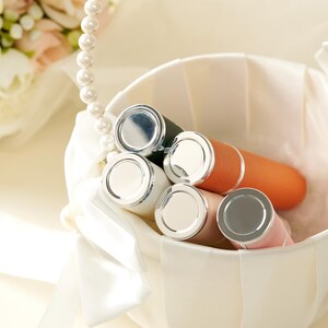May include: A collection of lipsticks in a white basket. The lipsticks have silver caps and various colored tubes, including white, orange, pink, and green. A pearl necklace and a bouquet of flowers are in the background.