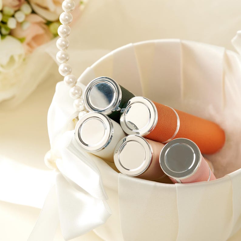 May include: A collection of lipsticks in a white basket. The lipsticks have silver caps and various colored tubes, including white, orange, pink, and green. A pearl necklace and a bouquet of flowers are in the background.