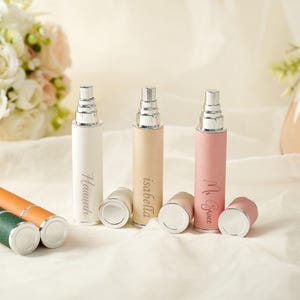 May include: Three personalized perfume atomizers in white, beige, and pink leatherette, each with a silver-tone spray nozzle and a matching cap. The names "Hannah", "Isabella", and "Ms. Grace" are engraved on the bottles. The background is a soft, white fabric.