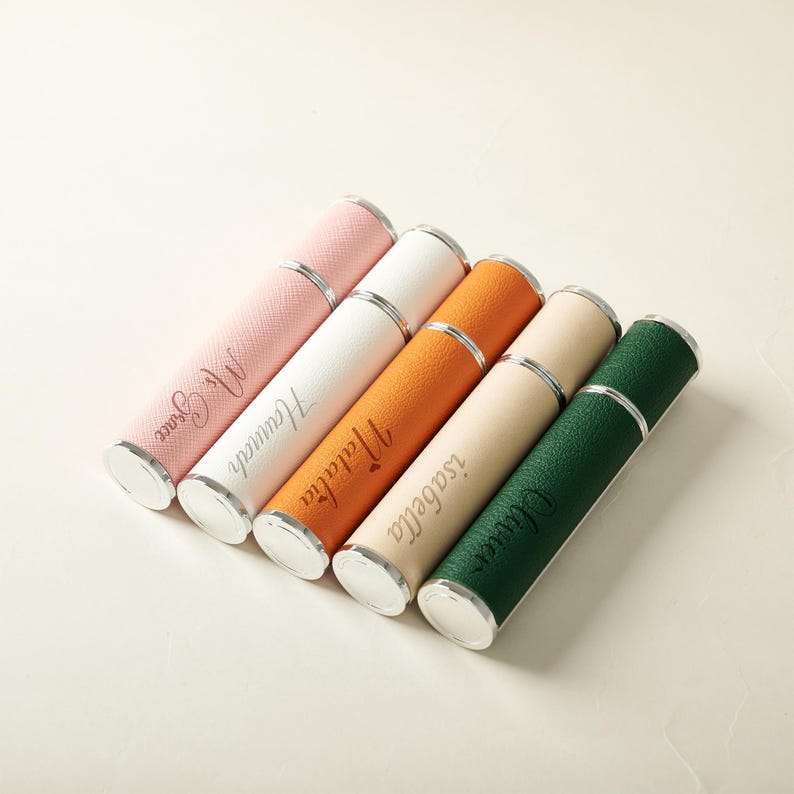 May include: A collection of six personalized perfume atomizers in various colors, including pink, white, orange, beige, and green. Each atomizer has a silver-colored band and base, with a name engraved on the side. The names are in a cursive font.