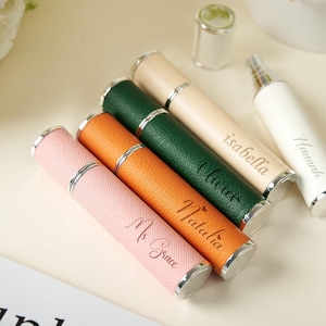May include: A collection of personalized perfume atomizers in various colors, including pink, orange, green, beige, and white. Each atomizer has a silver-colored band and cap, with names engraved on the side. The atomizers are cylindrical in shape.